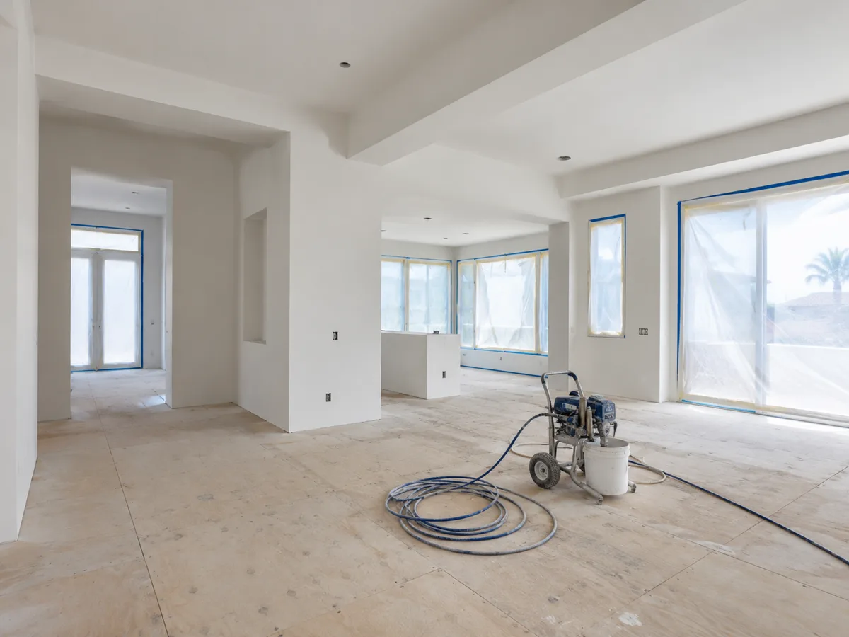 New-build interior with walls and ceiling sprayed flat white, windows masked with plastic and blue tape, a bare plywood subfloor and an airless paint sprayer with its hose coiled on the floor.